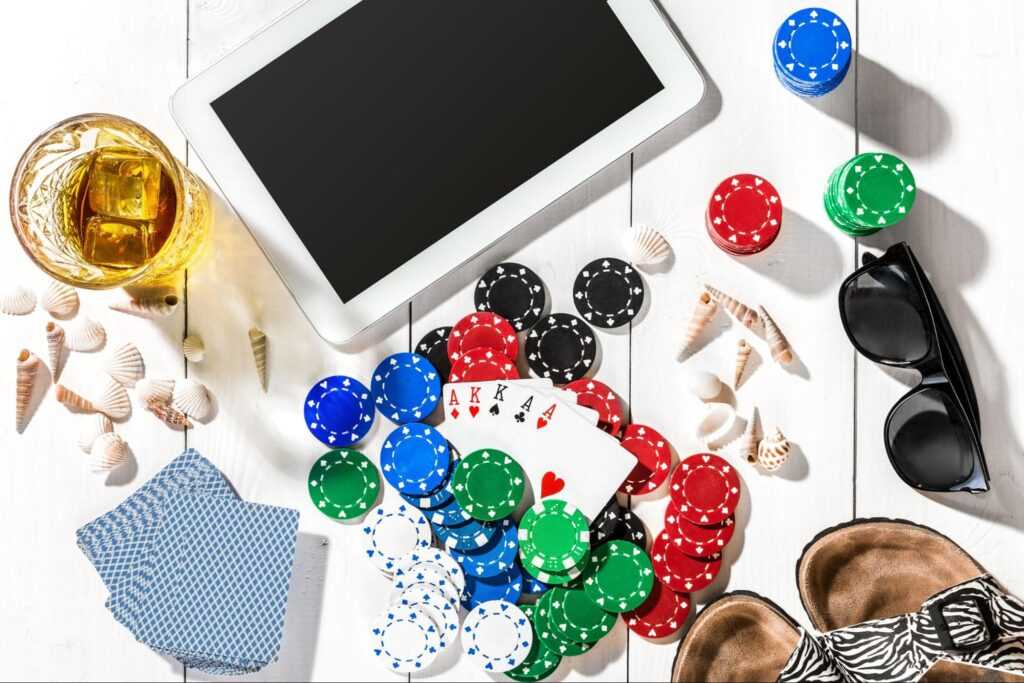 The image shows a top-down view of a wooden table with gambling items, including poker chips in various colors, playing cards, a tablet, a glass, seashells, sunglasses, and a pair of sandals, creating a summer-themed gambling setup.